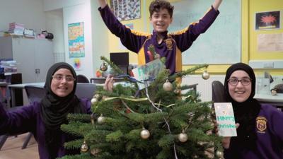 Three children stood around a Christmas tree with their arms in the air, looking happy.