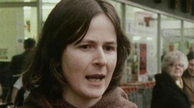 A woman with brown hair speaking on the street, in front of a shop window.