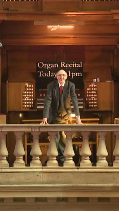 Organist stands on stone balcony in front of Kelvingrove organ.