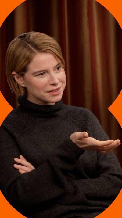 Jessie Buckley wears a dark grey jumper and sits with her arms crossed