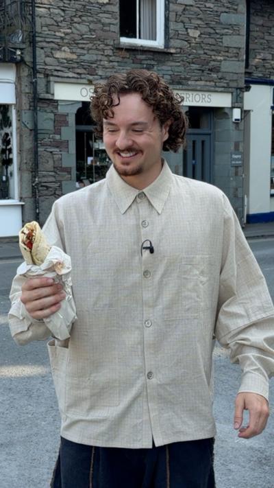 A person with curly hair and a light beige button-up shirt smiles while holding a wrapped kebab on a street in Grasmere, with traditional stone buildings in the background
