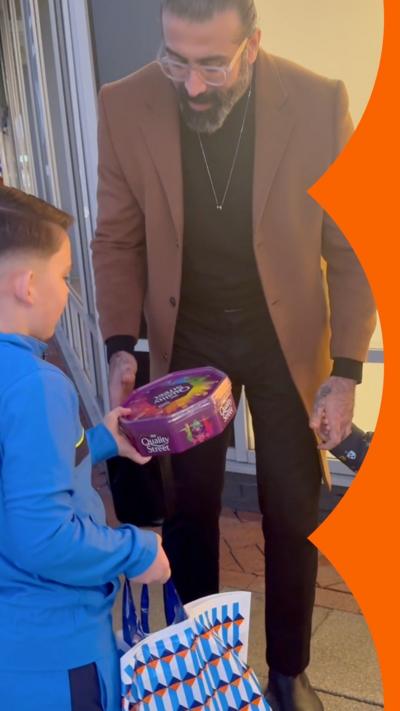 A boy in a blue tracksuit hands a box of chocolates to a man in the street