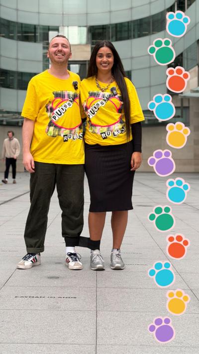 Two individuals stand together in a plaza, wearing bright yellow Children in Need branded t-shirts, surrounded by a busy urban environment. Their legs are tied together.
