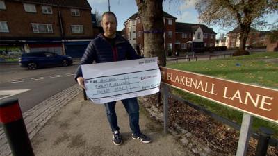 Man with a giant cheque stood next to Blakenall Lane road sign