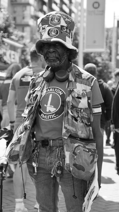 Norman Barker in camouflage attire and a Charlton Athletic t-shirt stands among a crowd outside a sports venue.