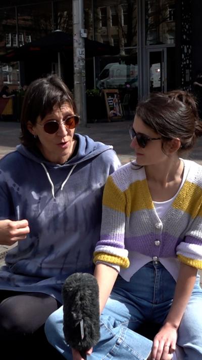 Two people sit on a seat along a pedestrian street. One is wearing a blue tie-dye hooded top and sunglasses, and the other is wearing a white, yellow and lilac stripe cardigan and sunglasses.