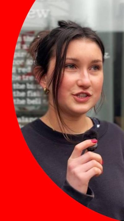 A girl with her mouth slightly open looks off to the side. She has dark hair that is tied up and hazel eyes. She is wearing gold earrings and a dark jumper. She stands in front of a glass window that has text on it.