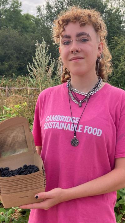 Alex Vardill, wearing a pink T-shirt with CAMBRIDGE SUSTAINABLE FOOD printed on it, stands outside, cradling a cardboard box containing blackberries. Alex looks at the camera. They have curly brown or ginger hair and wear various necklaces.