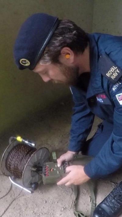 A picture of a Royal Navy officer crouched down holding a remote. He is in the corner of a room.