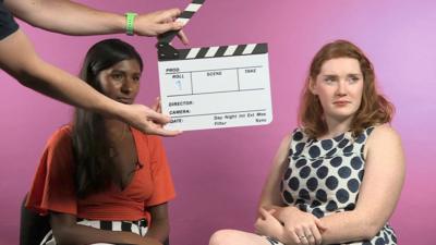 Two women wait seated, while a clapper board is held in front of them