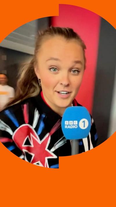 JoJo Siwa looks at the camera holding a Radio 1 microphone. She's wearing a brightly coloured jacket.
