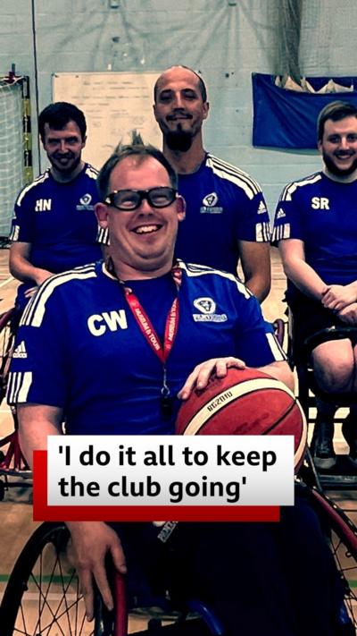 A wheelchair basketball team are wearing blue uniform and smiling. The team are gathered in a sports hall which has painted blue brick walls. Chris Wilson is in the foreground and has a red lanyard and whistle and a basketball on his lap.