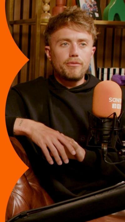 Roman Kemp wears a black hoody and talks into an orange BBC branded microphone