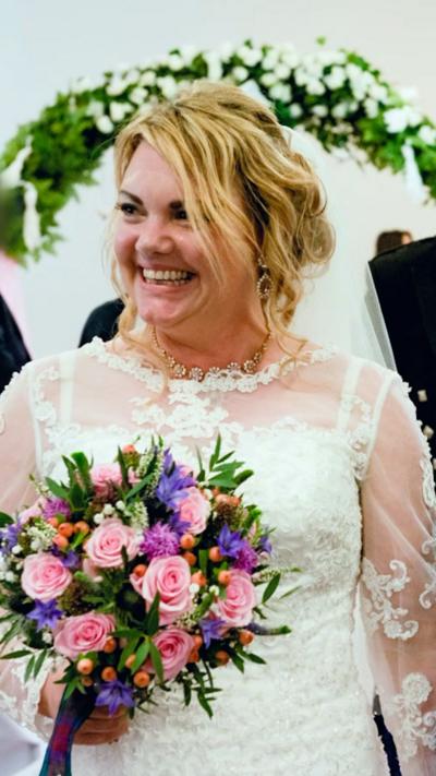A blonde woman wearing a white wedding dress and holding pink and purple flowers smiles off to the side. Behind her is a white wall and a garland of white flowers.