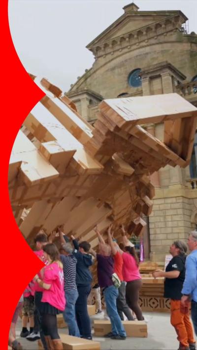 Group of people lifting up a large cardboard structure of a wheel with a triangle point at the top.
