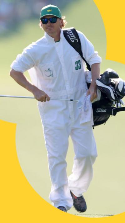 A Masters caddie wears a white jumpsuit with the number 33 on their left pocket. They wear a green cap and hold a bag of golf clubs.