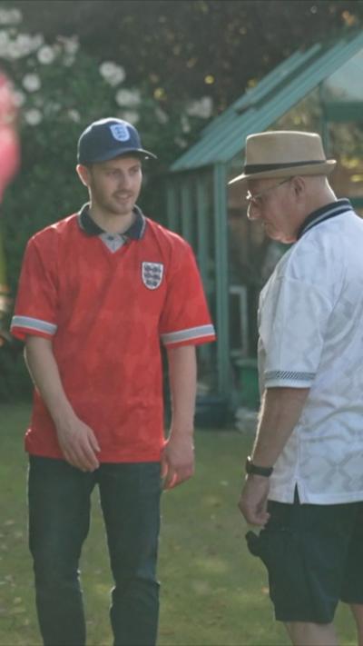 Two men stand chatting on a lawn. One wears a red England football shirt and blue cap, the other a white shirt, shorts, and a hat. Behind them is a garden and small greenhouse. A blurred flowering bush is to the left of them.