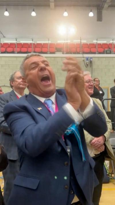 A man clapping his hands. He is wearing a blue suit. There are people in the background. 