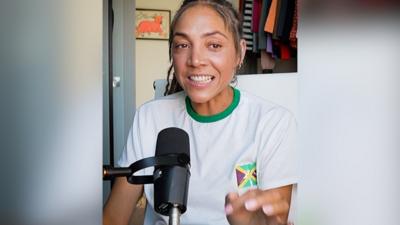 A woman, Miquita Oliver, wears a white T-shirt with the Jamaica flag and has long brown hair. She sits behind a microphone