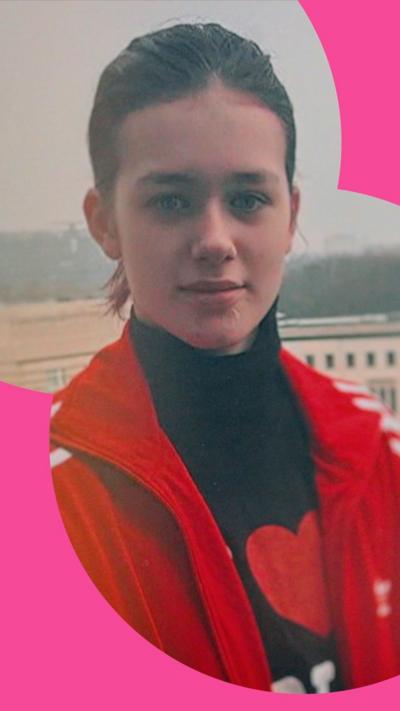A girl with pulled back brown hair  is wearing a high-necked black top with red and white decoration on it, and a red jacket.
