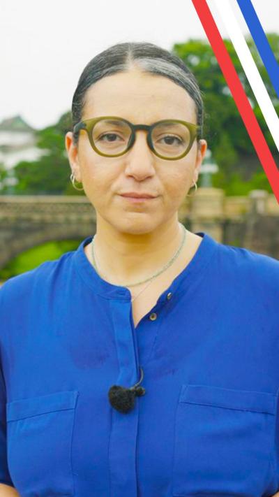 BBC Japan correspondent Shaimaa Khalil looks thoughtfully into the camera while stood in front of the Japanese Imperial Palace. Behind her is a grassy background with a stone bridge. She is wearing her hair back, round glasses and a blue shirt with a microphone clipped on to it.