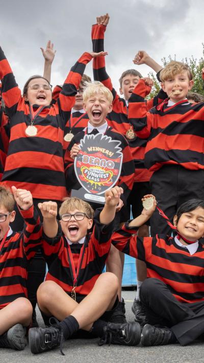 Rows of Year 6 pupils in matching red and black t-shirts modelled on Dennis the Menace. They are also wearing medals and have their hands in the air, cheering.