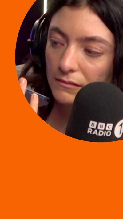 Lorde wearing headphones and sitting in front of a Radio 1 mic with her phone in her hand