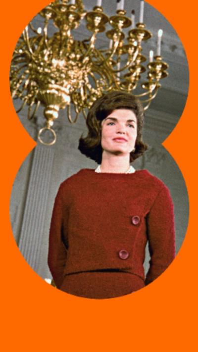 Jackie Kennedy wearing a red dress stand beneath a gold chandelier