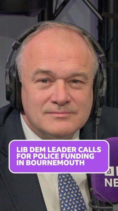 Ed davey wears headphones and looks at the camera, he has balding grey hair and wears a white shirt, blue and white patterned tie and a dark blue blazer.
