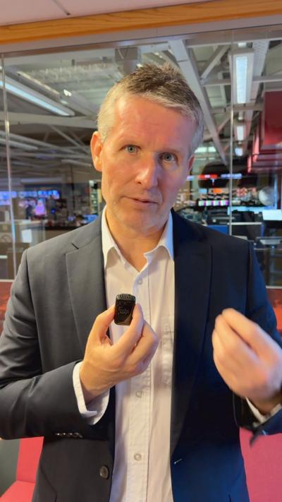 a man in a suit inside a newsroom with screens behind him. He is holding a small microphone and gesticulating