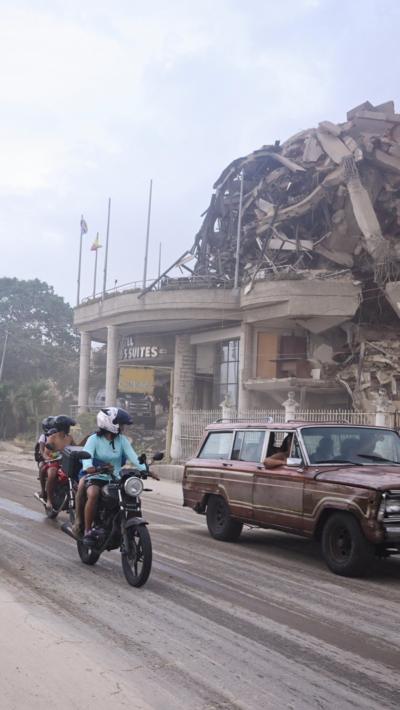 Image shows the aftermath of a hotel collapse. There is a car and two motorbikes in front of the building, which is collapsed in a pile in the background. 