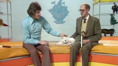 Peter Purves pats a white cat, sitting next to a man in a suit with a cane.