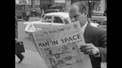 Man wearing suit reads the Evening Times on the side of the street. Headline on newspaper reads: MAN IN SPACE.