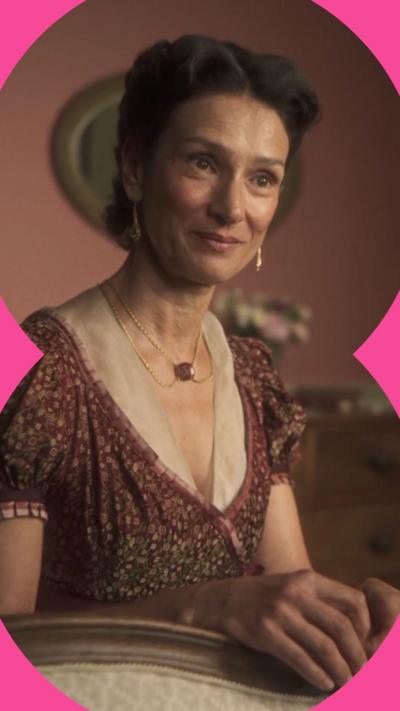 A woman with dark hair done in an elaborate hairdo is smiling. She wears a dark red patterned dress with a lace neckline.