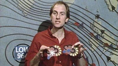 Blue Peter presenter Simon Groom, standing in front of an old-fashioned weather chart, holding some magnetic weather symbols.