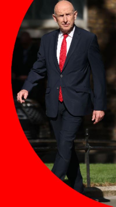 John Healey walking down Downing Street wearing a suit with a red tie.