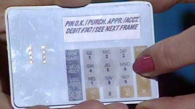 Closeup of a prototype of a future credit cards.  A finger and painted red fingernail of the hand holding the card can be seen on the right hand side.  Text on the card reads "PIN O.K. / PURCH. APPR. / ACCT. (next line) DEBIT #747 / SEE NEXT FRAME".  Below that there is a blue column of 4 buttons which say: "VISA CARD; CHECK ACCT; SAVE ACCT & CORP CARD".  There is also a numberpad and function buttoms similar to that on a phone number pad or calculator. 