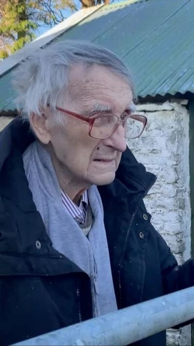 An old man with white hair and red-rimmed glasses. He is wearing a dark coat and grey jumper. He is staring to the right of the picture.Behind him is a white house with a green tin roof.