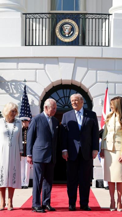 King Charles and President Donald Trump, both in blue suits, stand side-by-side outside the White House standing on a red carpet. The Queen and First Lady are by their sides