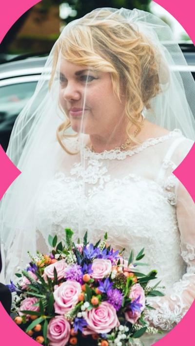 Charmain Speirs is wearing a white wedding dress with a veil over her head. She's stood in front of a car and is holding a bouquet of pink, purple, and orange flowers.