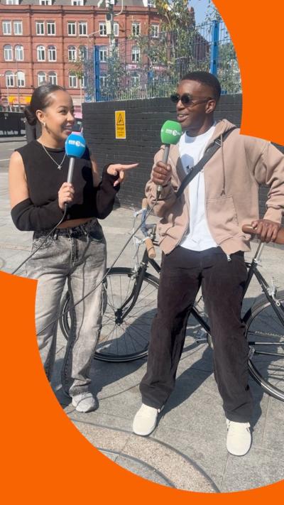 Kaylee Golding, holding a 1Xtra microphone, interviews people outside shops on a street.
