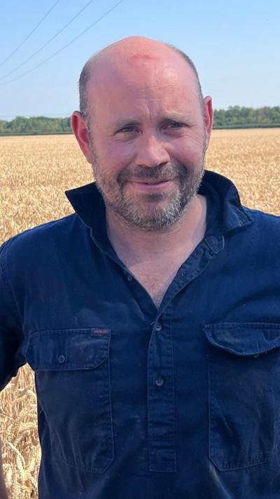A man with his hands on his hips wearing a denim shirt and he is smiling at the camera. Crops are behind him