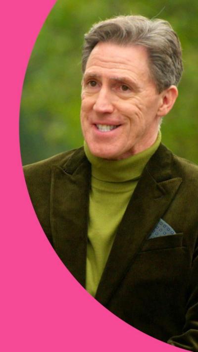 Rob Brydon smiling, wearing green polo neck and green blazer, in front of trees.