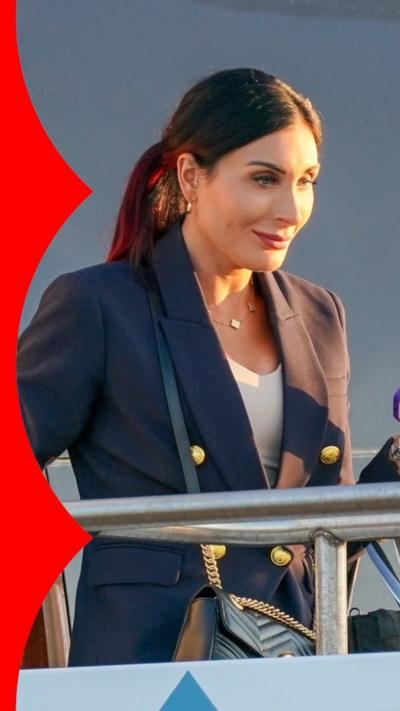 A woman with long dark hair steps off a plane carrying a phone and wearing a blue blazer.