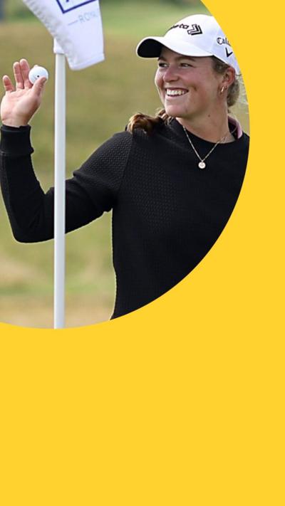 Golfer Mimi Rhodes smiles holding up a golf ball on the course - she wears a white cap and black top 