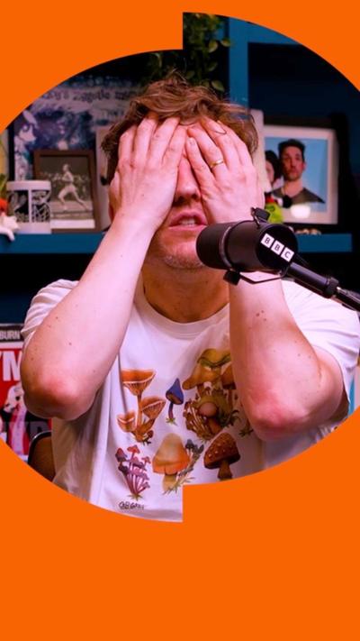 A man holds his hands over his eyes and head, in front of a mic. Behind him are blue shelves filled with posters and bric a brac.