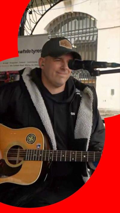 Man smiles while playing the guitar in the street. He is standing with a microphone in the foreground, in front of his face