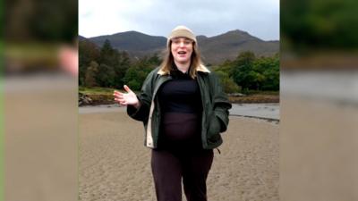 Elle Duffy - pregnant woman in dark clothing, a green jacket and tan hat, walking on a beach under a cloudy sky