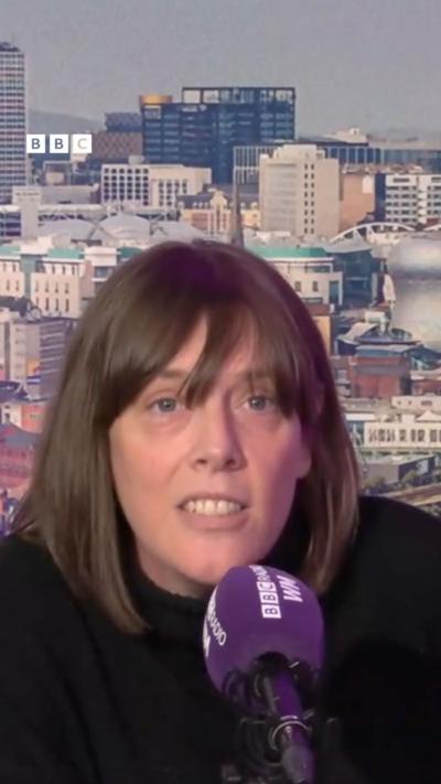 A woman with long brown hair talking into a purple microphone with the Birmingham skyline behind