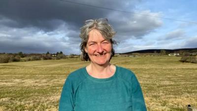 Fiona NicLeòid a tha an sàs sa bhuidhean Gàidhlig an Cataibh agus i na seasamh air a' bhlàr a-muigh 's gàire air a h-aodann.